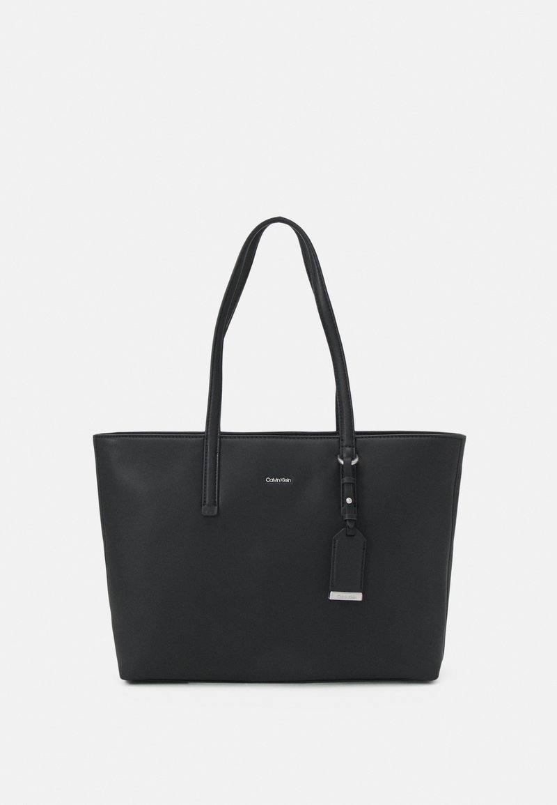 Sac cabas en cuir noir avec double poignée, logo Calvin Klein et étiquette suspendue attachée sur un fond uni.