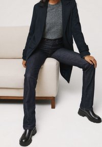 Una persona è seduta su un divano chiaro, indossando un cappotto scuro, un maglione grigio e jeans blu, con stivali neri su uno sfondo bianco.