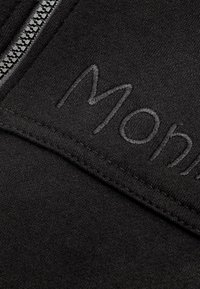 Zwarte sweatshirtstof met een close-up van een rits en geborduurd "Mon" detail in bijpassend garen, dat de gladde textuur en stiksels toont.