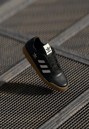 Czarny sneaker Adidas z białymi paskami i gumową podeszwą unoszący się nad metalową kratą w naturalnym świetle.