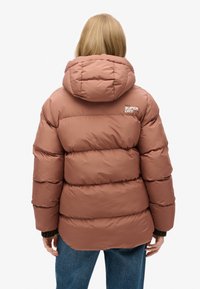 Veste doudoune rose avec un design matelassé et une capuche. Présente le logo "Superdry" dans le haut du dos. Le matériau semble lisse et isolant.