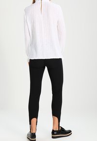 Blusa bianca a maniche lunghe con texture e apertura a chiave sul retro, abbinata a pantaloni neri slim-fit con spacchi laterali e scarpe nere.