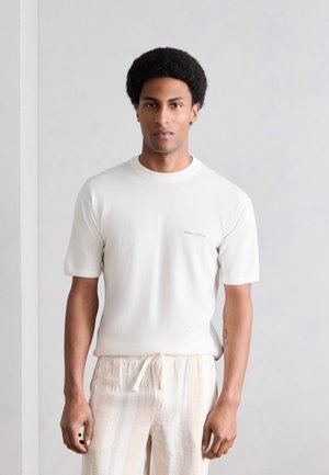 Jeune homme avec coiffure afro portant une chemise blanche à manches courtes et un pantalon beige clair rayé, debout devant un fond clair uni.