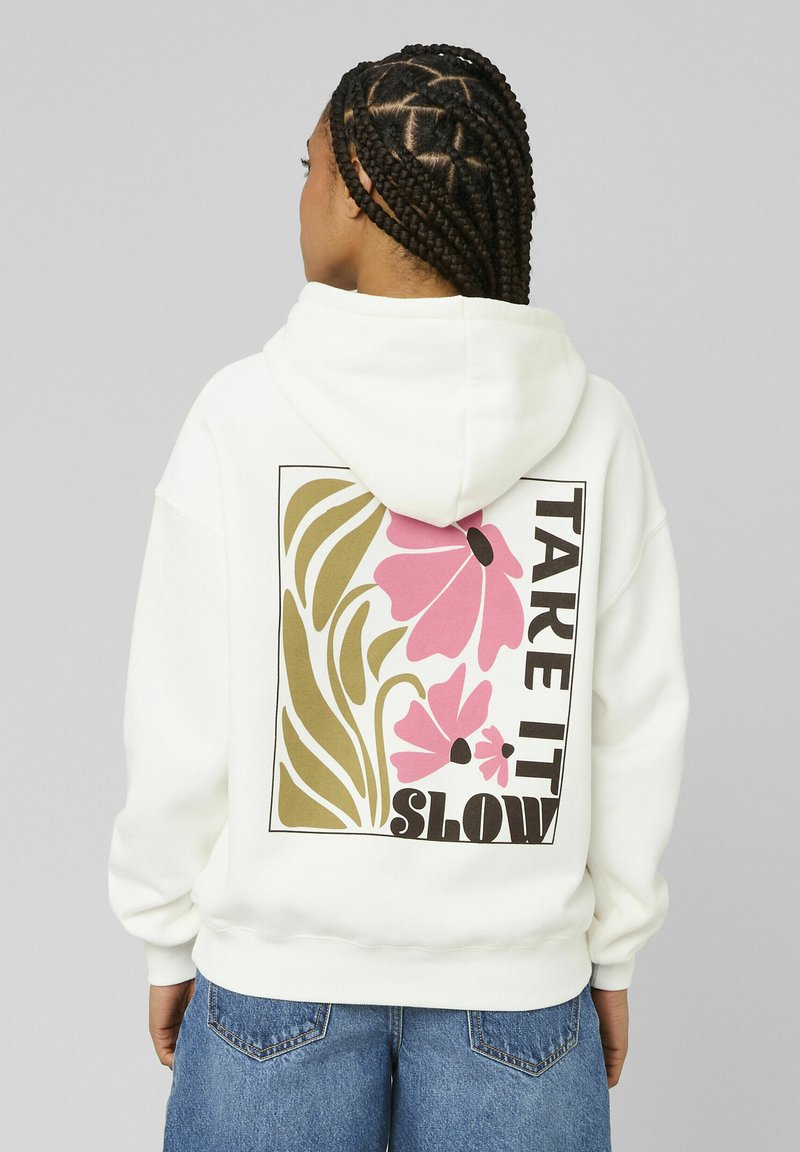 Personne aux cheveux tressés portant un sweat à capuche blanc orné de fleurs roses et le texte "TAKE IT SLOW" dans le dos, associée à un jean bleu.