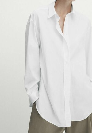 Weit geschnittenes weißes Hemd mit Knöpfen und langen Ärmeln, getragen über beigen Hosen, gezeigt vom Brustbereich bis zu den oberen Oberschenkeln.