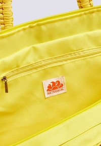 Borsa a spalla in tessuto giallo con tasca con zip e un'etichetta bianca con un logo arancione. Manici testurizzati con motivo intrecciato.