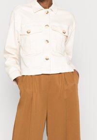 Cream-colored cropped jacket with gold-tone buttons, two chest pockets, and a collared neckline, paired with brown wide-leg trousers.