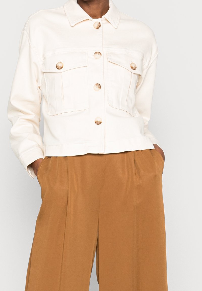Cream-colored cropped jacket with gold-tone buttons, two chest pockets, and a collared neckline, paired with brown wide-leg trousers.
