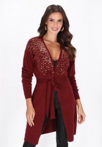 Mosolygó nő, bordó hosszú kardigánt visel, amelynek vállán flitterek vannak, és megkötött övvel rendelkezik, fekete nadrággal párosítva.