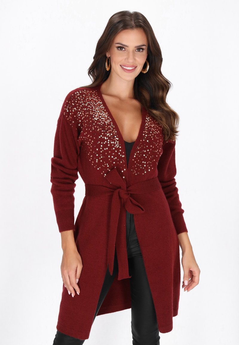 Mosolygó nő, bordó hosszú kardigánt visel, amelynek vállán flitterek vannak, és megkötött övvel rendelkezik, fekete nadrággal párosítva.