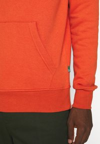 Orange sweatshirt med känguruficka, ribbat ärmslut och en figurnära nederkant. Materialet verkar mjukt med en strukturerad yta.