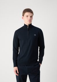 Navy zip-up sweater made of soft cotton. Features a high collar, ribbed cuffs, and a small embroidered logo on the chest.