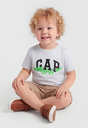 Batole s kučeravými blond vlasmi, sediace na zemi so skríženými nohami, oblečené v bielom tričku GAP s grafikou zeleného krokodíla a v béžových šortkách s hnedými topánkami.