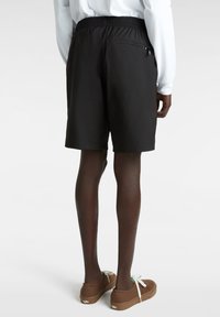 Schwarze Shorts mit einem elastischen Bund, ausgestattet mit einer Reißverschluss-Tasche auf der Rückseite und einem glatten, leichten Material, kombiniert mit braunen Sneakers.