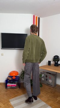 Zelen pulover, sive široke hlače, brušeni rjavi čevlji. Pohištvo z oranžnimi in vijoličnimi poudarki. Preprost dizajn, teksturirani materiali, notranja postavitev.