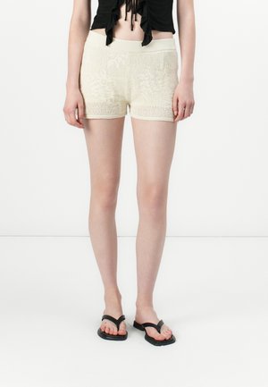 Knit cream shorts with a textured pattern, featuring an elastic waistband and a snug fit. Paired with black flip-flops.