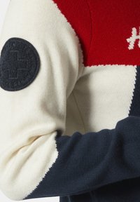 Suéter de punto con bloques de color rojo, crema y azul marino. Destacada parche redondo negro en la manga con emblema. Textura suave y diseño ajustado.