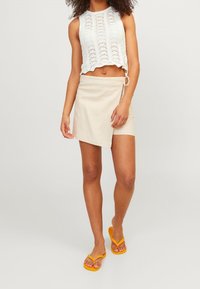Vit spetscroptop med scallopkant, i kombination med en beige omslagskjol. Modellen har på sig gula flip-flops. Enkel, avslappnad sommarmundering.