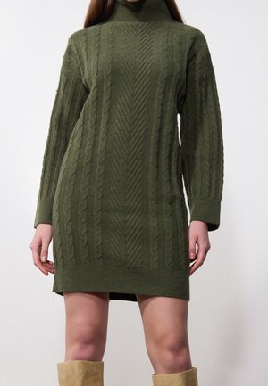 Kobieta ubrana w oliwkowozieloną, grubą, dzianą sukienkę z golfem i długimi rękawami oraz beżowe kozaki za kolano.