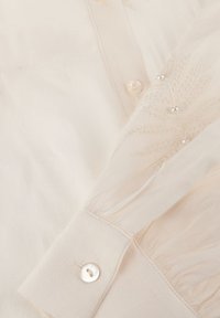 Close-up van de mouw van een crèmekleurige blouse met een knop, doorschijnende gerimpelde mouw en delicate parelversieringen.