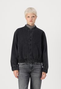 Svart kort denimjacka med ståkrage, elastisk nederkant och ärmslut samt två framfickor. Modellen bär en grå topp och jeans.