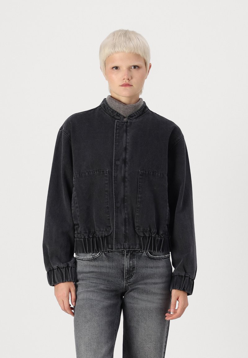 Svart kort denimjacka med ståkrage, elastisk nederkant och ärmslut samt två framfickor. Modellen bär en grå topp och jeans.