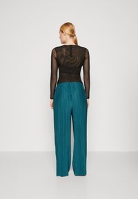 Sort gennemsigtig bluse med lange ærmer, parret med plisserede teal bukser med bred ben. Bukserne har en sammenfoldet talje og lodrette plisseringer for struktur.