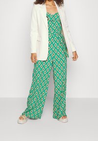 Žena v zeleném overalovém kostýmu s geometrickým vzorem a uvázaným pasem, bílém blejzru a béžových platformových sandálech na pozadí jednoduché bílé stěny.