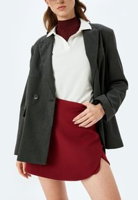 Grauer Blazer über einem burgunderfarbenen Rock und einem weißen Polo. Der Blazer hat einen Brustknopf, eine glatte Textur, einen scharfen Kragen und eine taillierte Form.