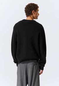 Homme aux cheveux bouclés portant un pull noir et un pantalon gris à fines rayures, vu de dos sur un fond clair uni.