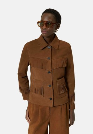 Kobieta w brązowej zamszowej kurtce z frędzlami na piersi, kieszeniach i mankietach, w zestawie z dopasowanymi spodniami i okularami przeciwsłonecznymi.