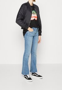 Veste bomber noire, t-shirt graphique noir avec un motif de dents colorées, jean évasé bleu clair, et baskets Vans noires avec des détails blancs.