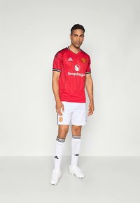 Camisola de futebol vermelha do Manchester United com detalhes em preto, calções brancos e meias com riscas pretas. Usada com chuteiras brancas.