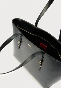 Sac fourre-tout en cuir noir avec des accents de métal doré, comprenant un compartiment intérieur zippé et une étiquette de marque rouge. Deux bretelles solides pour l'épaule.