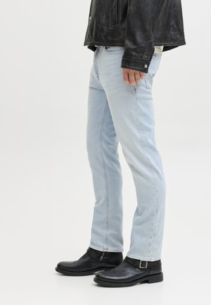 Persona valkā gaiši zilas džinsa bikses, melnu ādas jaku un melnas potīšu zābaciņus ar sprādzēm, stāvot pret vienkāršu baltu fona sienu.