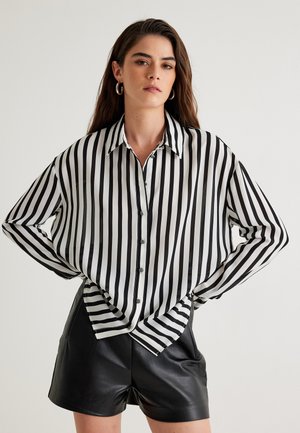 Gestreifte Schwarz-Weiß-Hemdbluse mit Kragen und langen Ärmeln, kombiniert mit schwarzen Leder-Shorts. Weicher Stoff, lockere Passform.