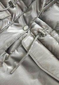 Tissu matelassé argenté brillant avec un bouton-pression en métal, un cordon de serrage avec un arrêt en métal, et une poche à rabat.