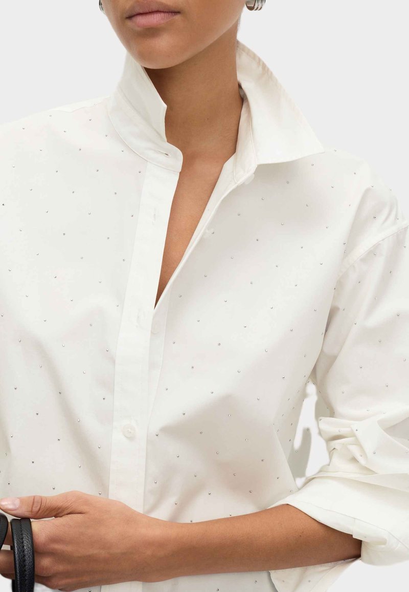 Mujer vistiendo una camisa blanca de botones con pequeños remaches plateados, con el cuello levantado y las mangas arremangadas, sosteniendo una correa de un bolso negro.