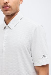 Άνδρας που φοράει λευκό πικέ πόλο με κουμπιά και το λογότυπο της Adidas στο αριστερό μανίκι, πάνω σε απλό λευκό φόντο.
