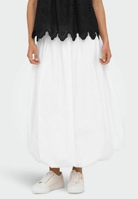 Svart broderad topp med vågig nederkant, kombinerad med en fladdrande vit kjol och beige sneakers, som framhäver lätta material och avslappnad stil.