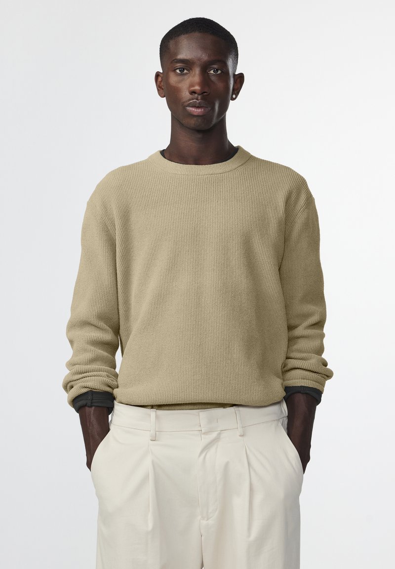Beige gebreide trui met een ronde hals en een ribbelstructuur, gecombineerd met crèmekleurige plooibroek. De stof lijkt zacht en heeft een ontspannen pasvorm.