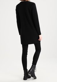 Černá tunika s dlouhým rukávem z texturované látky, s bočními kapsami a rovný střihem, kombinovaná s černými legínami a kotníkovými botami.