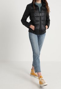 Svart, vadderad jacka med hög krage och dragkedjestängning, med horisontella quiltade sektioner. Bärs över en teal-färgad skjorta och avklippta jeans.