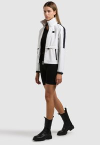 Femme portant une veste blanche zippée avec une bande noire, un short noir et des bottes noires mi-mollet, debout devant un fond uni.