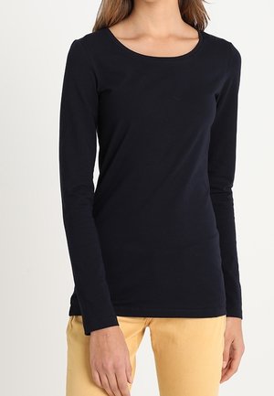 Kvinde iført en tætsiddende sort langærmet skjorte og lysegule bukser, stående mod en ensfarvet hvid baggrund.