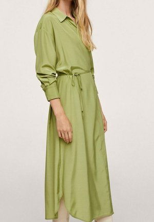 Femme portant une robe-chemise midi manches longues vert clair avec un col et une ceinture à la taille, se tenant devant un fond clair uni.