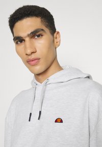 Sudadera de algodón gris con capucha ajustable y un pequeño logo bordado en naranja y rojo. Textura suave y corte relajado.