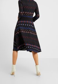 Vestido negro de mangas largas, con un patrón geométrico multicolor y un dobladillo acampanado, combinado con botas de tacón alto en color crema.