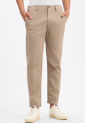 Personne portant un pantalon chino beige et des baskets blanches, les mains dans les poches, debout sur un fond blanc uni.