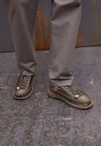 Beige woven sandals with thick soles, featuring an intricate lace pattern and white accent details, paired with gray checkered pants.